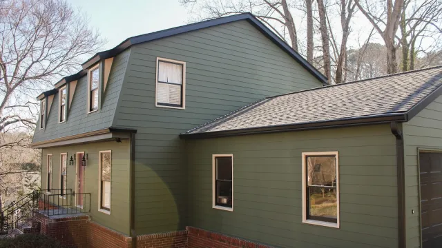 Two-story green house with brown trim, brick foundation, and a small front porch surrounded by trees in early spring.