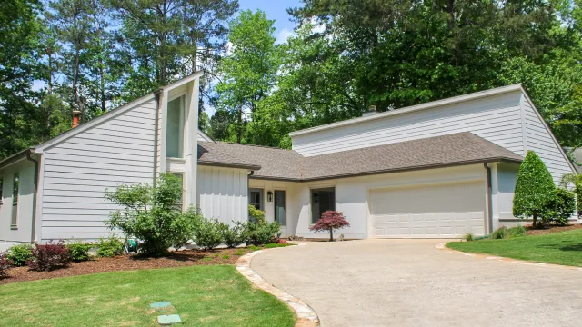 Modern single-story house with white siding, a two-car garage, and a curved driveway surrounded by greenery.