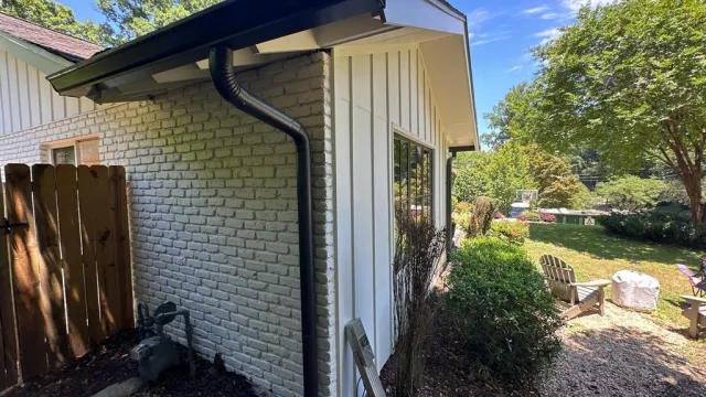 Side of white brick and siding house with black gutter, garden area, wooden fence, and sunny backyard with chairs and basketball hoop.