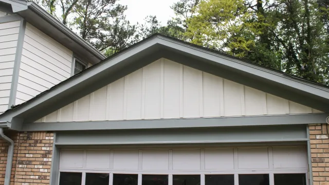 Residential garage door with white panels and windows under gray-trimmed gable roof surrounded by trees.