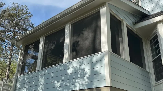 Modern two-story house with grey siding, multiple windows, wooden deck, and outdoor grill area on a sunny day.