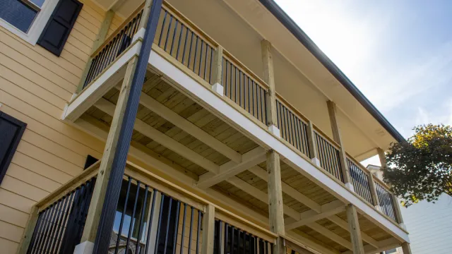 Two-story wooden balcony with black metal railings attached to light yellow house under a blue sky.