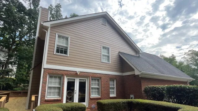 Two-story brick and beige siding house with white-trimmed windows and trimmed green bushes under a cloudy sky
