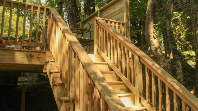 Wooden outdoor staircase with railings leading to a forest deck with chairs surrounded by trees and greenery.