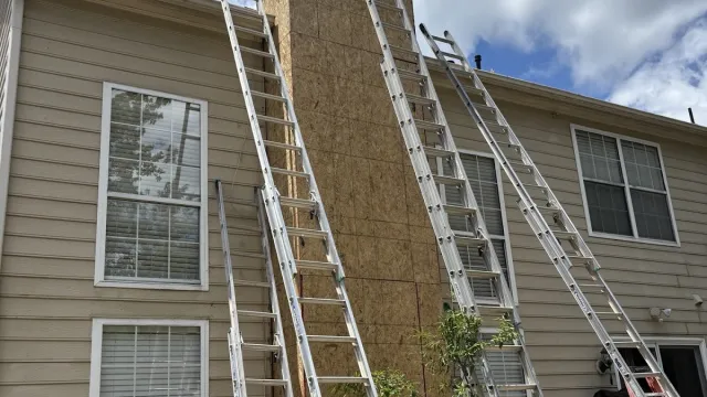 Multiple metal ladders leaning against a beige house chimney under a partly cloudy sky during construction.