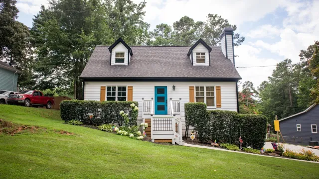 Charming white cottage with blue door, dormer windows, and manicured bushes on a green lawn under a cloudy sky.