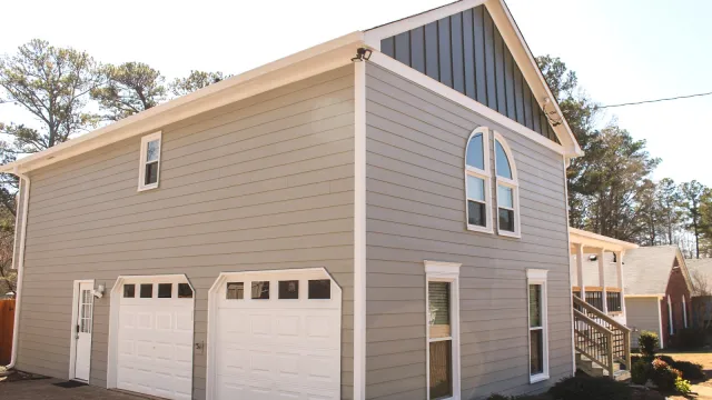 Two-story gray house with white-trimmed windows and double garage doors in sunny suburban setting