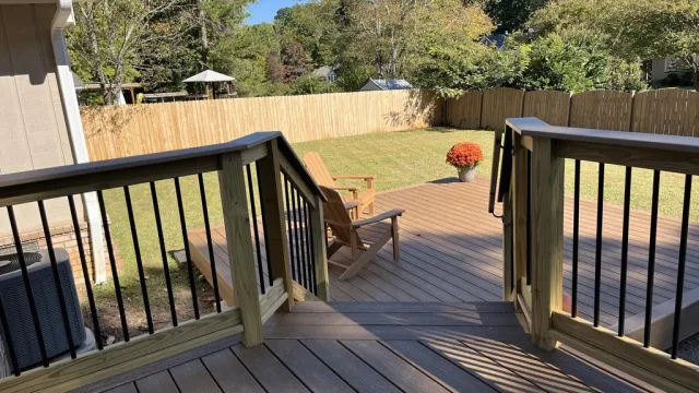 Wooden deck with railings, Adirondack chairs, potted flowers, and a large fenced backyard under clear blue sky.