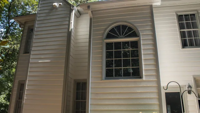 White two-story house with black shutters, glass front door, surrounded by green bushes and tall pine trees.