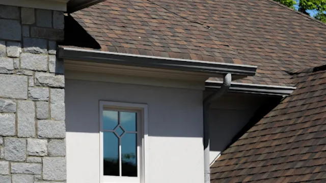 House exterior showing stone wall, white framed window, and brown shingled roof with gutter and blue sky.
