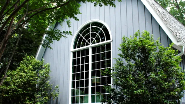 Gray house exterior with large arched window surrounded by lush green trees and manicured shrubs.