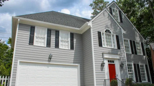 Two-story gray house with white trim, black shutters, red front door, and white double garage under blue sky.