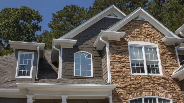 Modern two-story house with grey siding, multiple windows, wooden deck, and outdoor grill area on a sunny day.