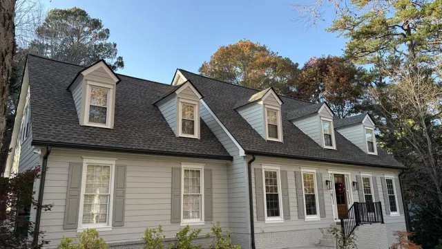 White two-story house with black roof, dormer windows, and surrounding autumn trees under blue sky