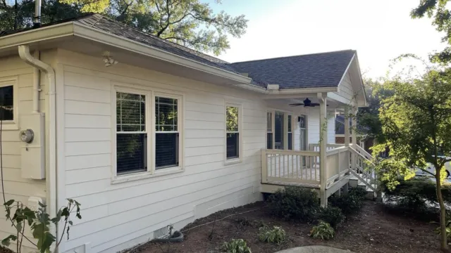 White single-story house with a porch surrounded by trees and a small garden under clear sky.