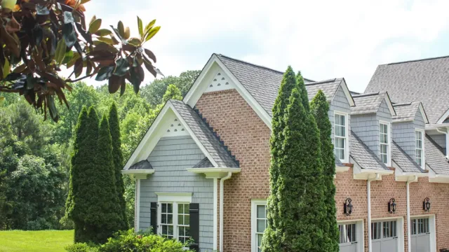 Suburban house with brick and gray siding, tall evergreen trees, and manicured green lawn on a sunny day