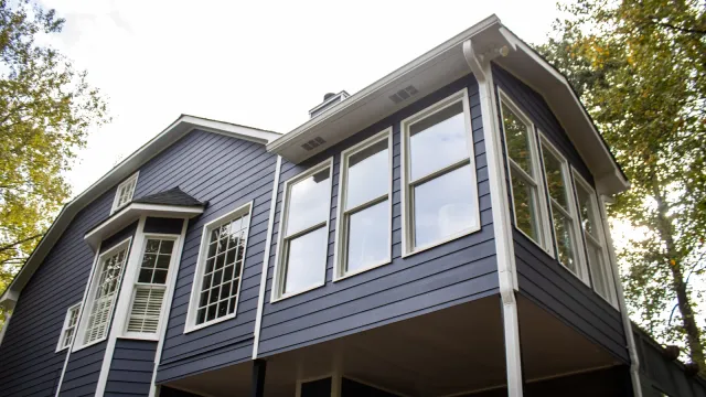 Modern two-story house with blue siding, large windows, and white trim surrounded by trees on a clear day.
