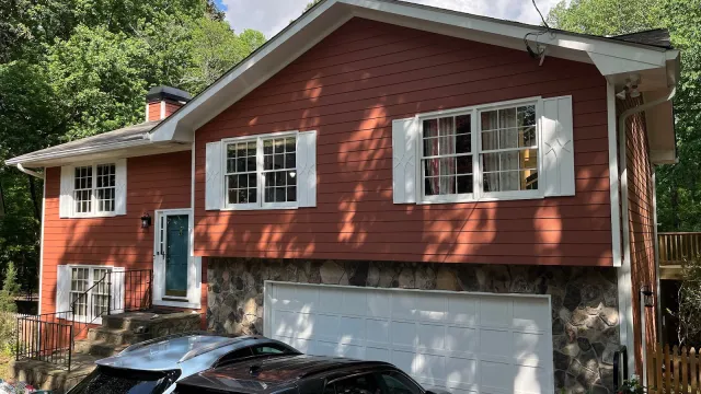 White two-story house with black shutters, glass front door, surrounded by green bushes and tall pine trees.