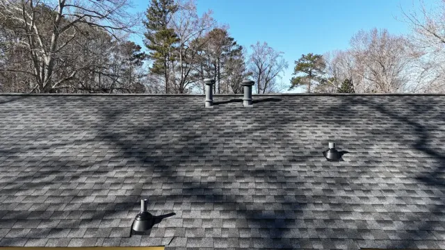 Gray asphalt shingle roof with three vent pipes and tree shadows under a clear blue sky.