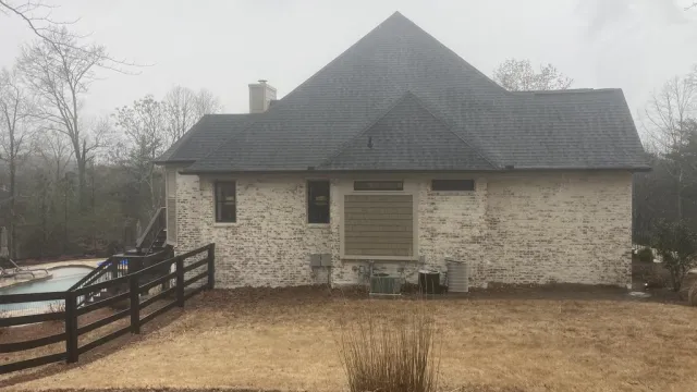 Back view of a brick house with black roof, fenced yard, dry grass, and a pool in a foggy setting.
