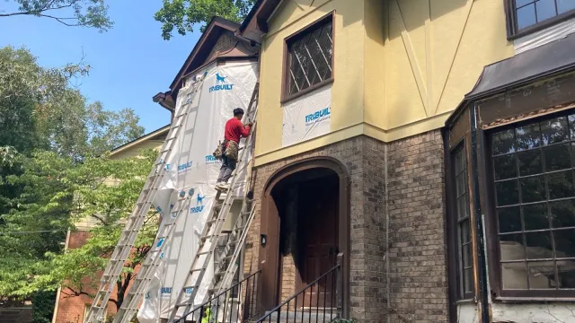 Worker on ladder installing siding on a two-story house exterior during daytime renovation.