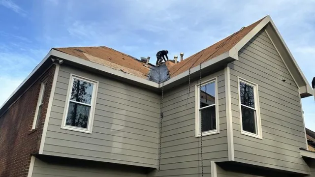 Worker installing shingles on roof of two-story modern house with gray siding and multiple windows under blue sky