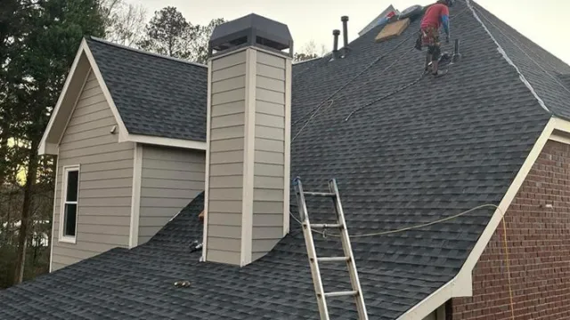 Roofer working on steep gray shingled roof with ladder and chimney during daytime near trees.