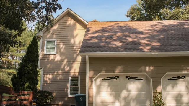 Suburban house exterior with beige siding, double white garage doors, and surrounding trees casting shadows.