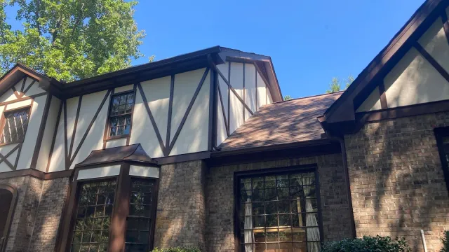 White two-story house with black roof, dormer windows, and surrounding autumn trees under blue sky