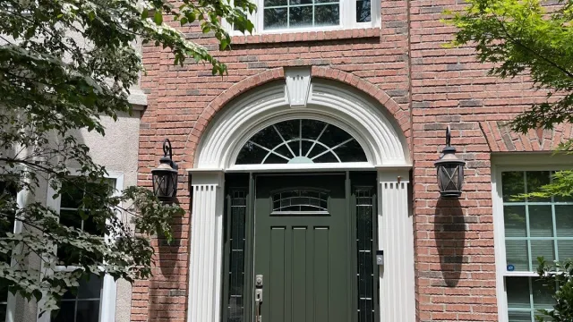 Brick house front with green door, arched white trim, sidelights, and wall lanterns surrounded by greenery.
