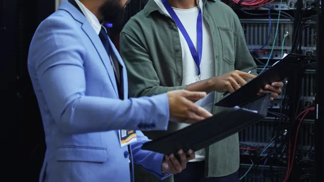 Two IT professionals discussing data on tablets in a server room filled with networking equipment.