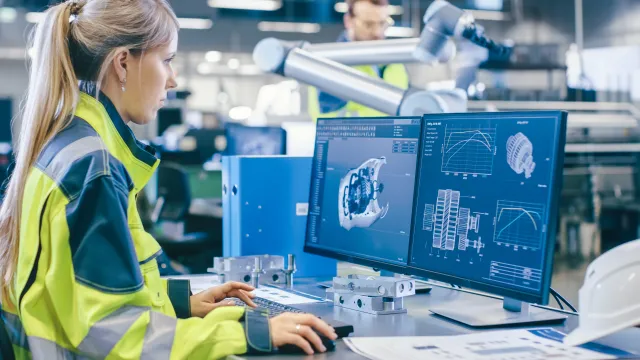 Engineer working on computer designs in a modern manufacturing facility with robotic arms in the background.