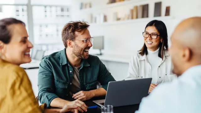 A diverse group of four people engaging in a lively discussion around a laptop in an office setting.
