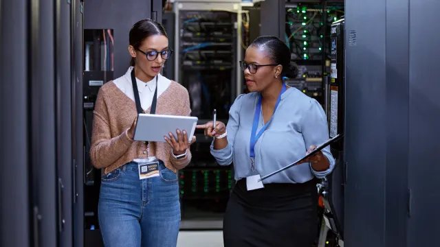 Two women discussing data in a server room, one using a tablet, the other holding a clipboard.