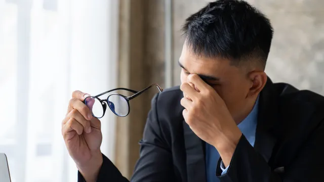 A stressed businessman adjusts his glasses while sitting at a desk with documents and a laptop.