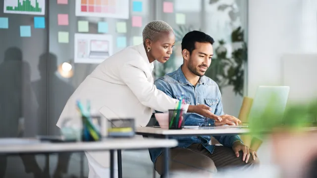 A businesswoman assisting a colleague with a laptop in a modern workspace.