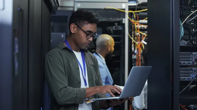 Man working on a laptop in a data center, surrounded by server equipment and wiring.