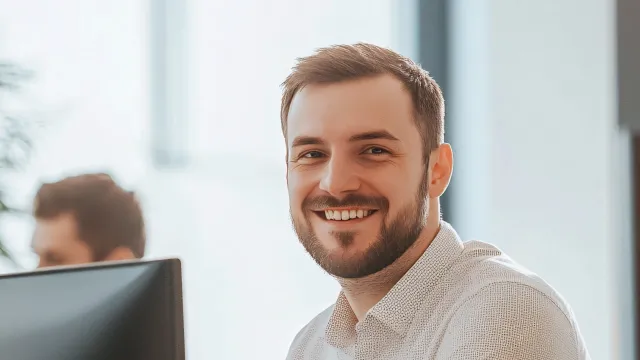 Smiling man at a desk in an office, working on a computer with colleagues in the background.