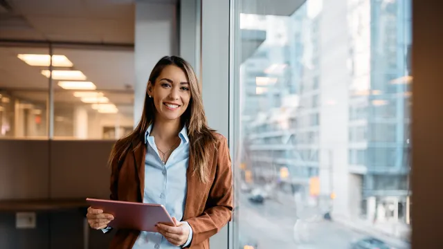 Smiling woman in a brown jacket holding a tablet, standing by an office window with a city view.