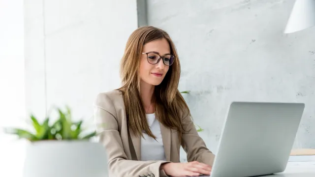 Professional woman working on a laptop in a modern office setting with a potted plant nearby.