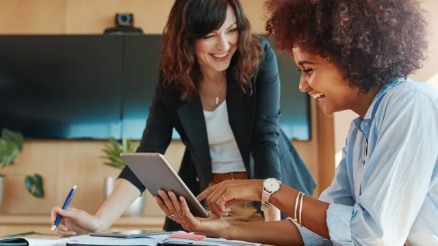 Two women collaborating in a modern office, using a tablet and discussing notes on a table.