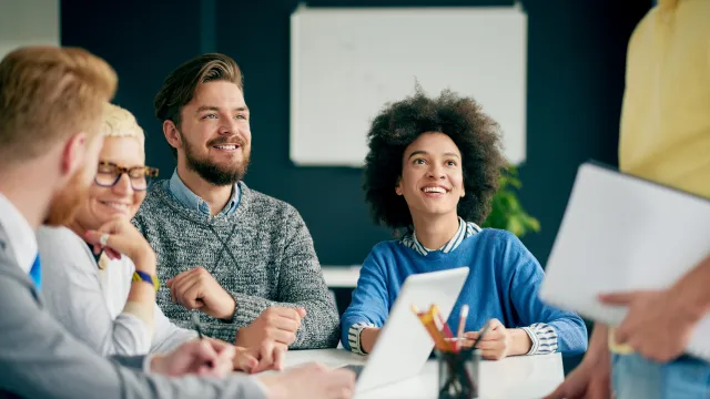 Diverse team in a meeting, smiling and engaged around a laptop and notepad on the table.