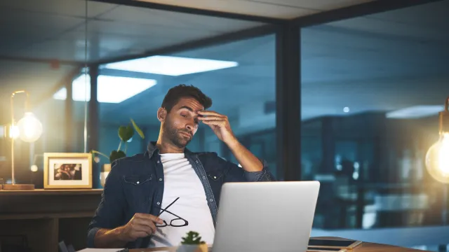 Frustrated man working late at desk with laptop, holding glasses in modern office setting.