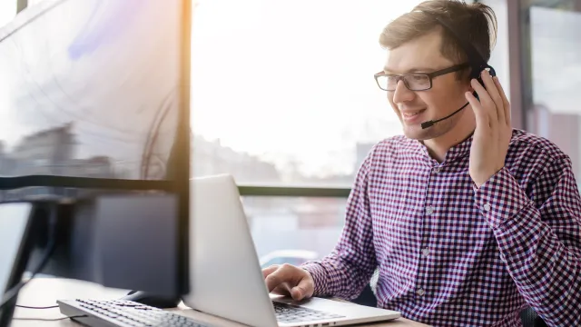 Man with headset working at a laptop, smiling in a bright office environment.