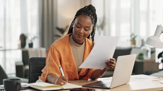 A woman working on a laptop, taking notes, in a bright, modern office space.