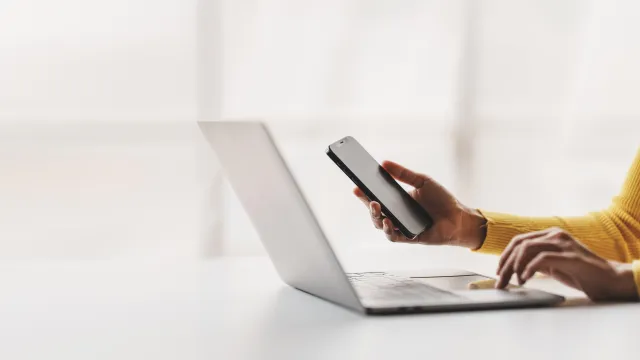 A person holding a smartphone while working on a laptop in a bright office setting.