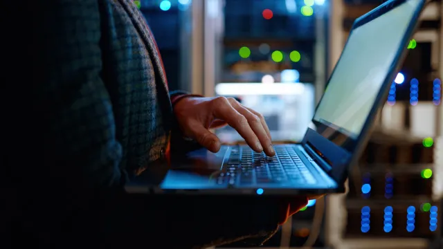 Person using a laptop in a server room with illuminated equipment in the background.