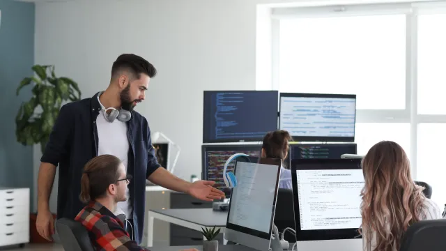 A team collaborating in an office, discussing work at computers with coding on screens and modern décor.
