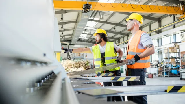 Two workers in hard hats and safety vests operating machinery in a bright industrial workshop.