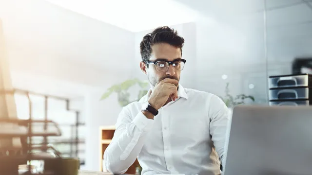 Man in glasses working intently on a laptop in a bright office setting with notebooks and a coffee mug.